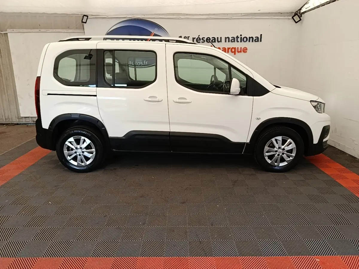 Profil droit d’un Peugeot Rifter blanc 2020 avec barres de toit chromées et jantes alliage en intérieur showroom.