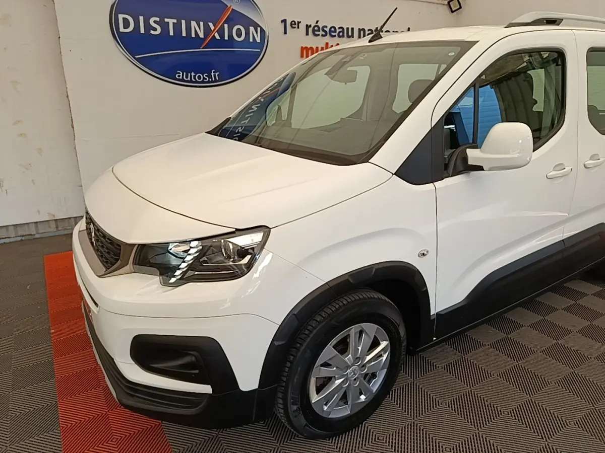 Vue 3/4 avant droit d'un Peugeot Rifter blanc avec phares LED et rétroviseurs blancs dans un showroom.
