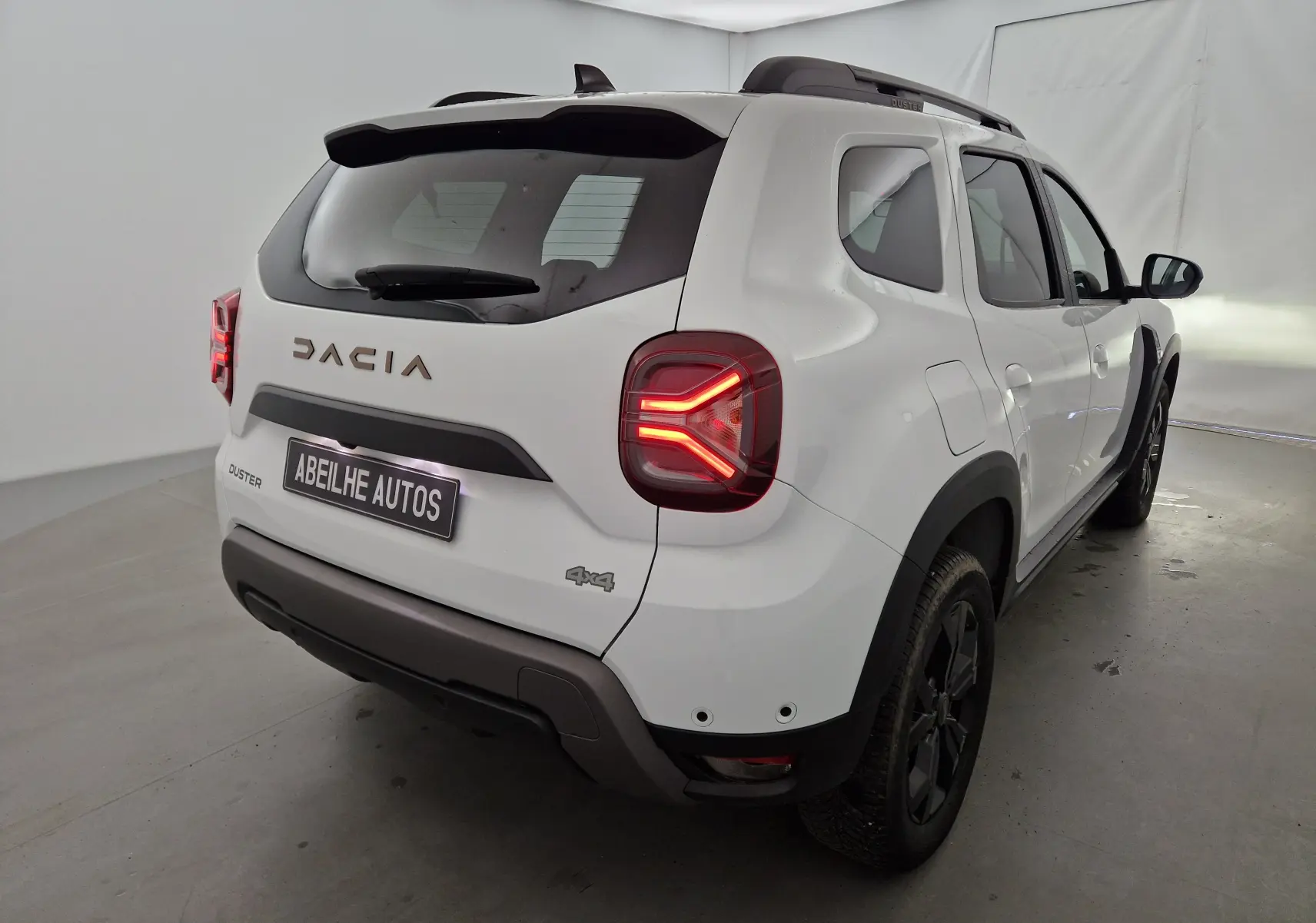 Vue 3/4 arrière droite du Dacia Duster blanc 4x4 avec feux arrière allumés et jantes noires distinctives.