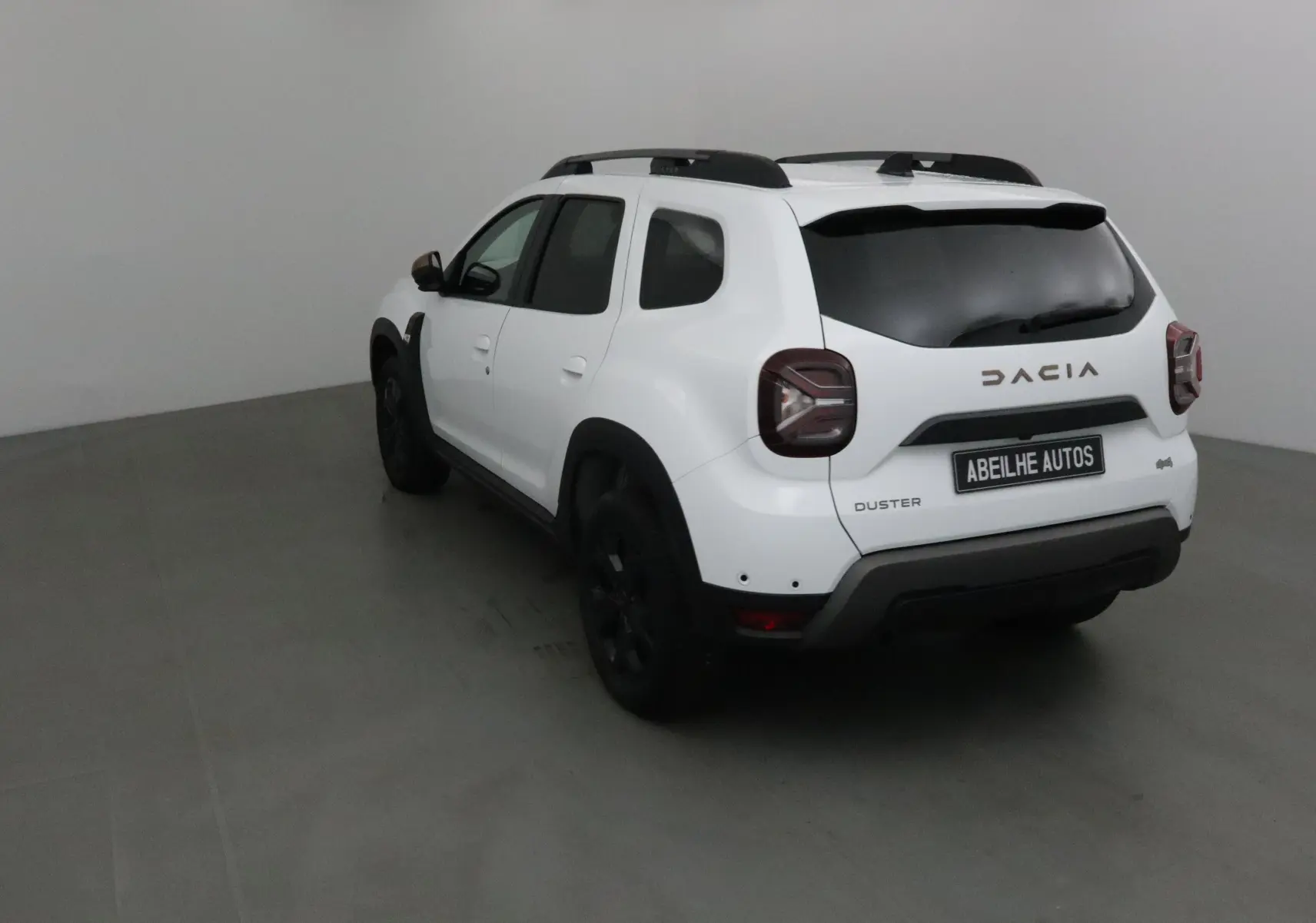 Vue 3/4 arrière gauche d'un Dacia Duster blanc 2024 avec barres de toit et jantes noires dans un studio.