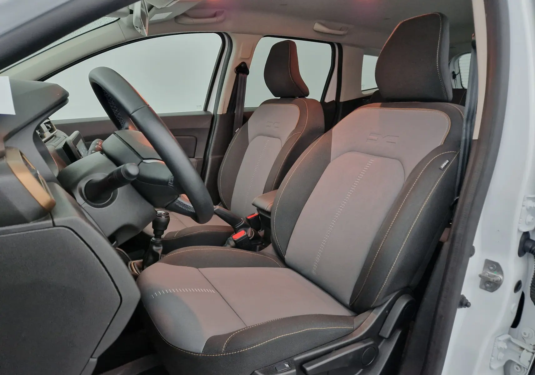 Intérieur du Dacia Duster 4x4 blanc 2024 vu côté gauche, sièges avant gris avec surpiqûres contrastées et volant visible.