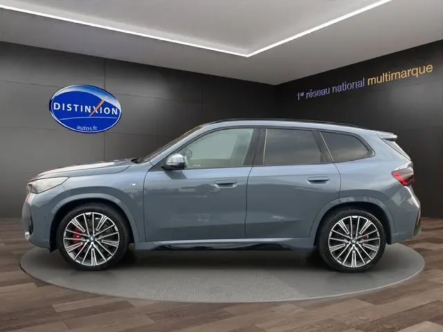 Vue latérale gauche d'un BMW X1 Storm Bay avec jantes alliage 20'' et freins M Sport rouges dans un showroom.