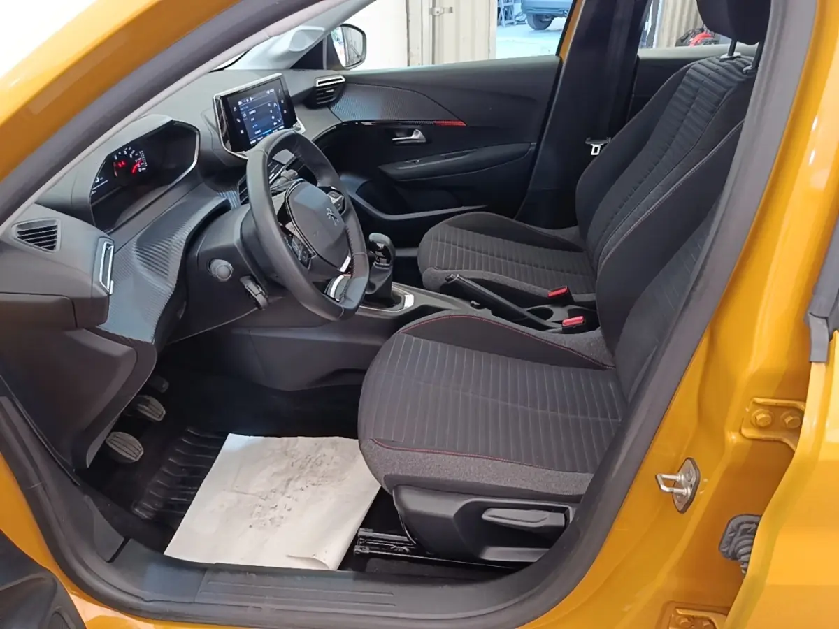 Vue intérieure côté conducteur de la Peugeot 208 jaune 2023, volant cuir multifonctions et sièges tissu gris à surpiqûres rouges.