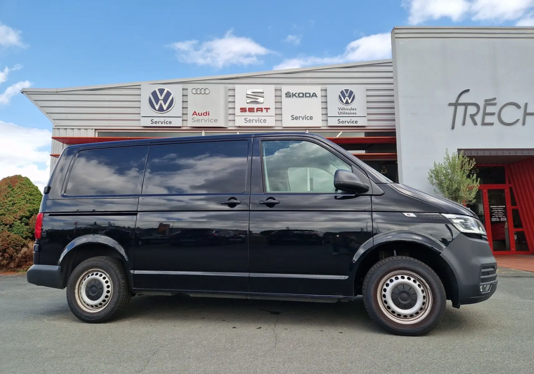 Profil côté droit d'un Volkswagen Transporter noir 2020 garé devant un centre de service multimarques.