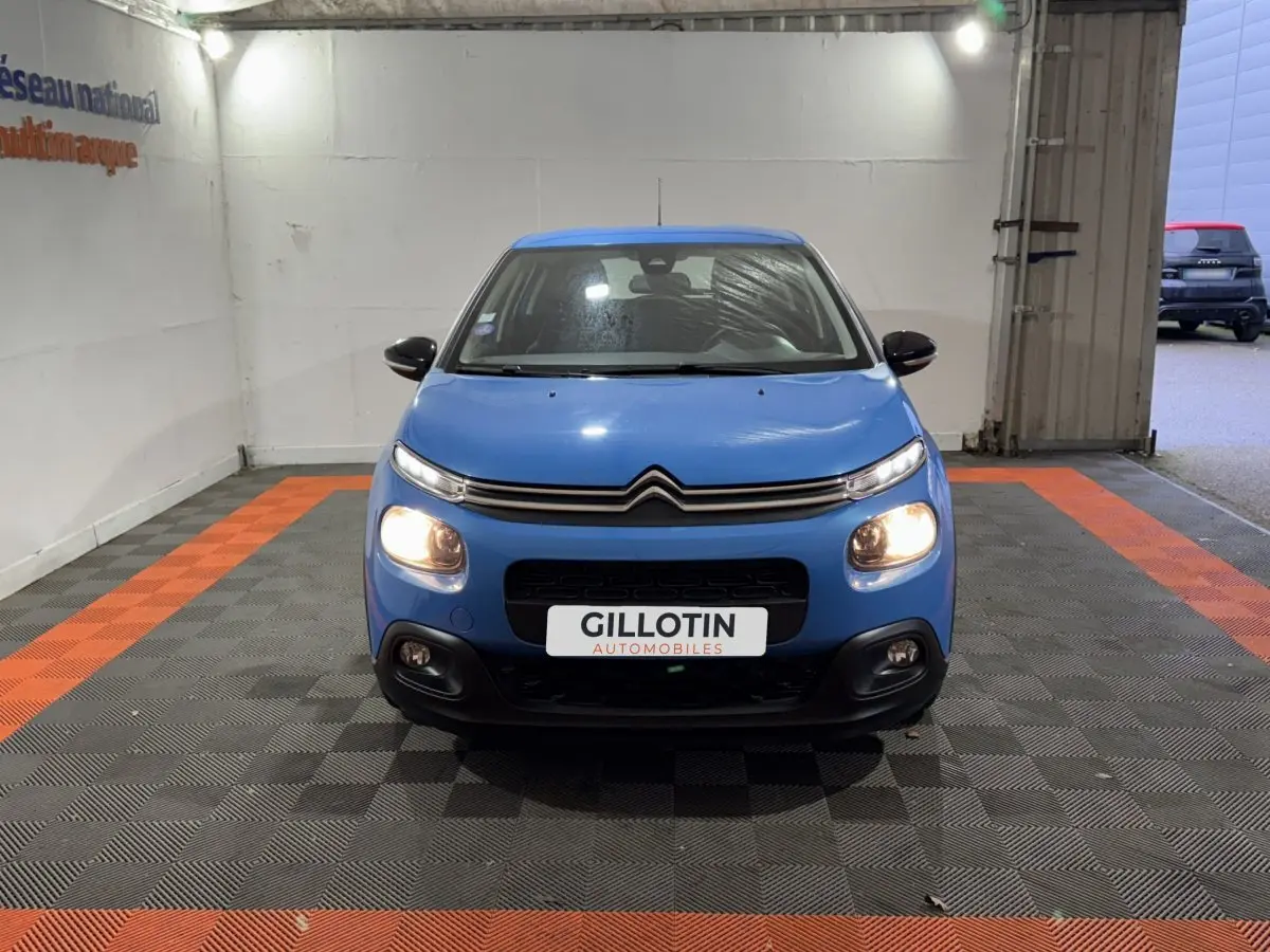 Citroën C3 bleu vue de face en intérieur avec phares allumés et logo distinctif sur calandre.