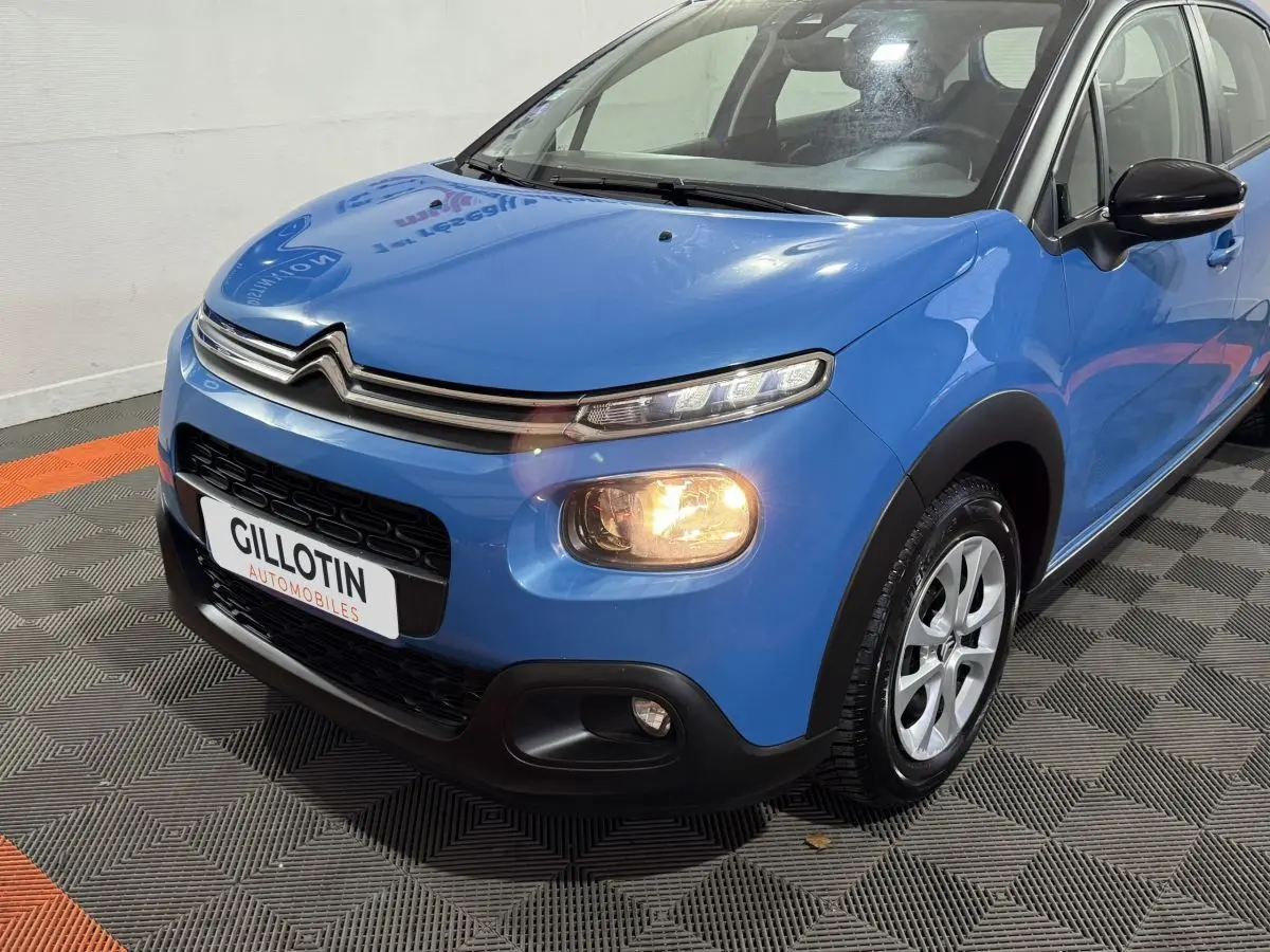 Vue 3/4 avant droite d'une Citroën C3 bleu avec phares allumés et calandre chromée distinctive.