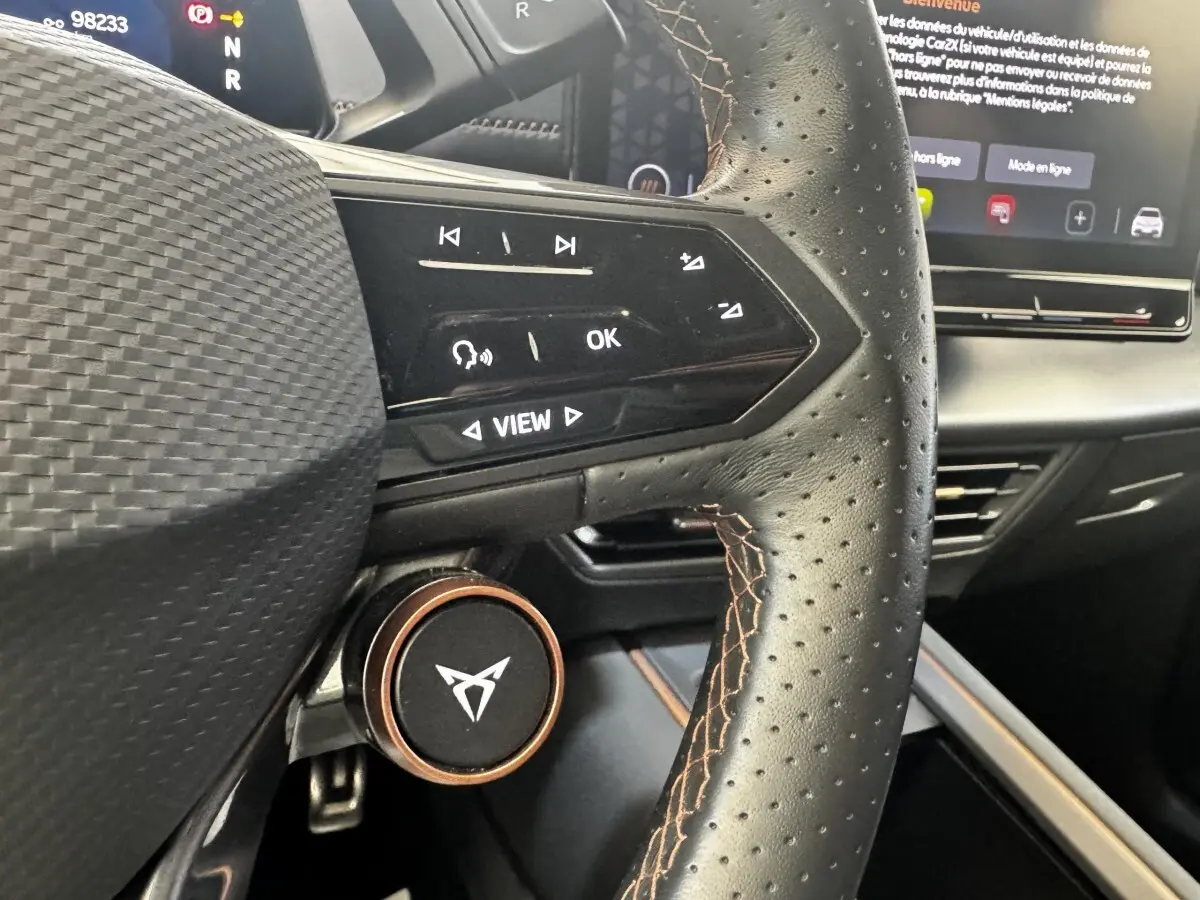 Gros plan sur le volant cuir perforé avec commandes tactiles et logo CUPRA, tableau de bord et écran tactile en arrière-plan.