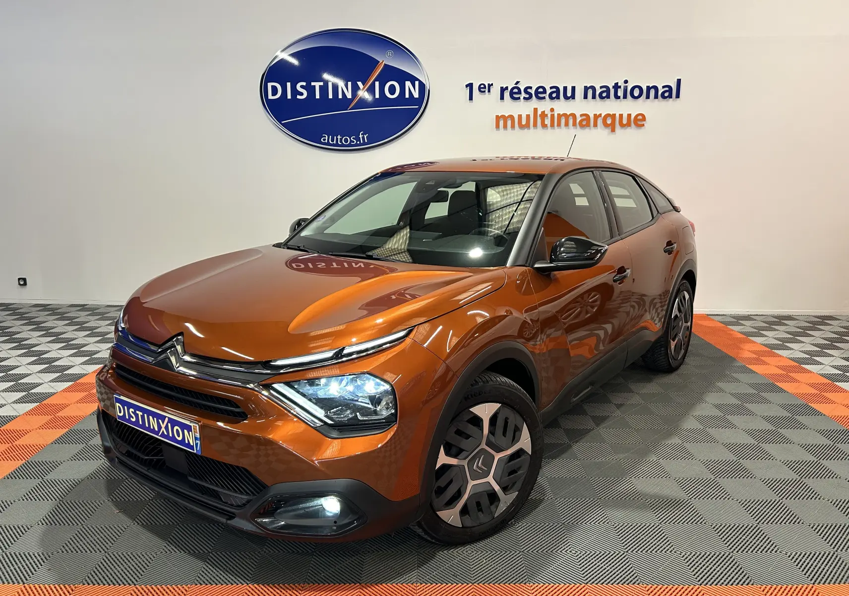 Citroën C4 PureTech 100 orange vue 3/4 avant droit avec feux allumés et attelage visible