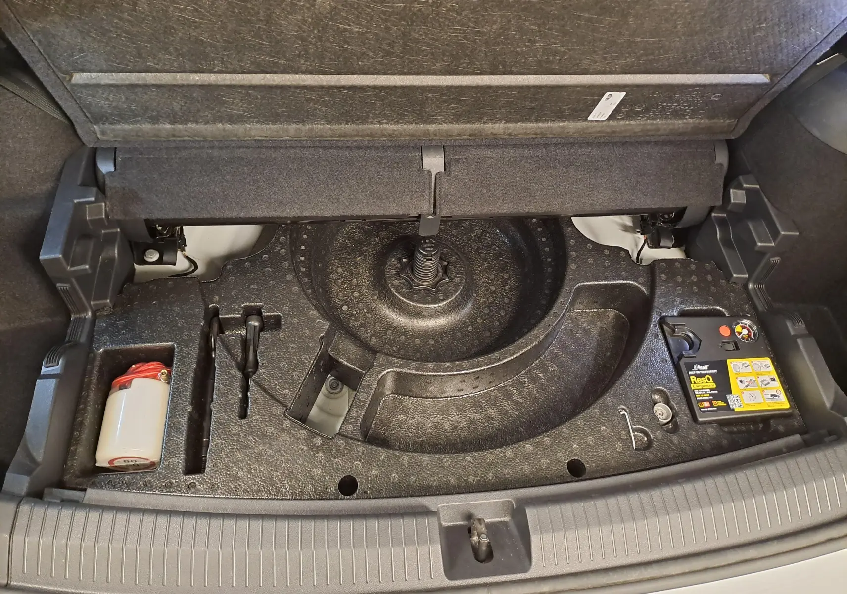 Coffre ouvert du Volkswagen Tiguan Allspace blanc, vue plongeante sur le rangement sous plancher avec kit de réparation pneus.