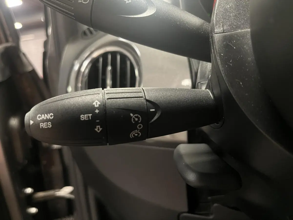 Gros plan sur la commande de régulateur de vitesse noire à gauche du volant dans l'habitacle d'une Fiat 500 gris 2023.