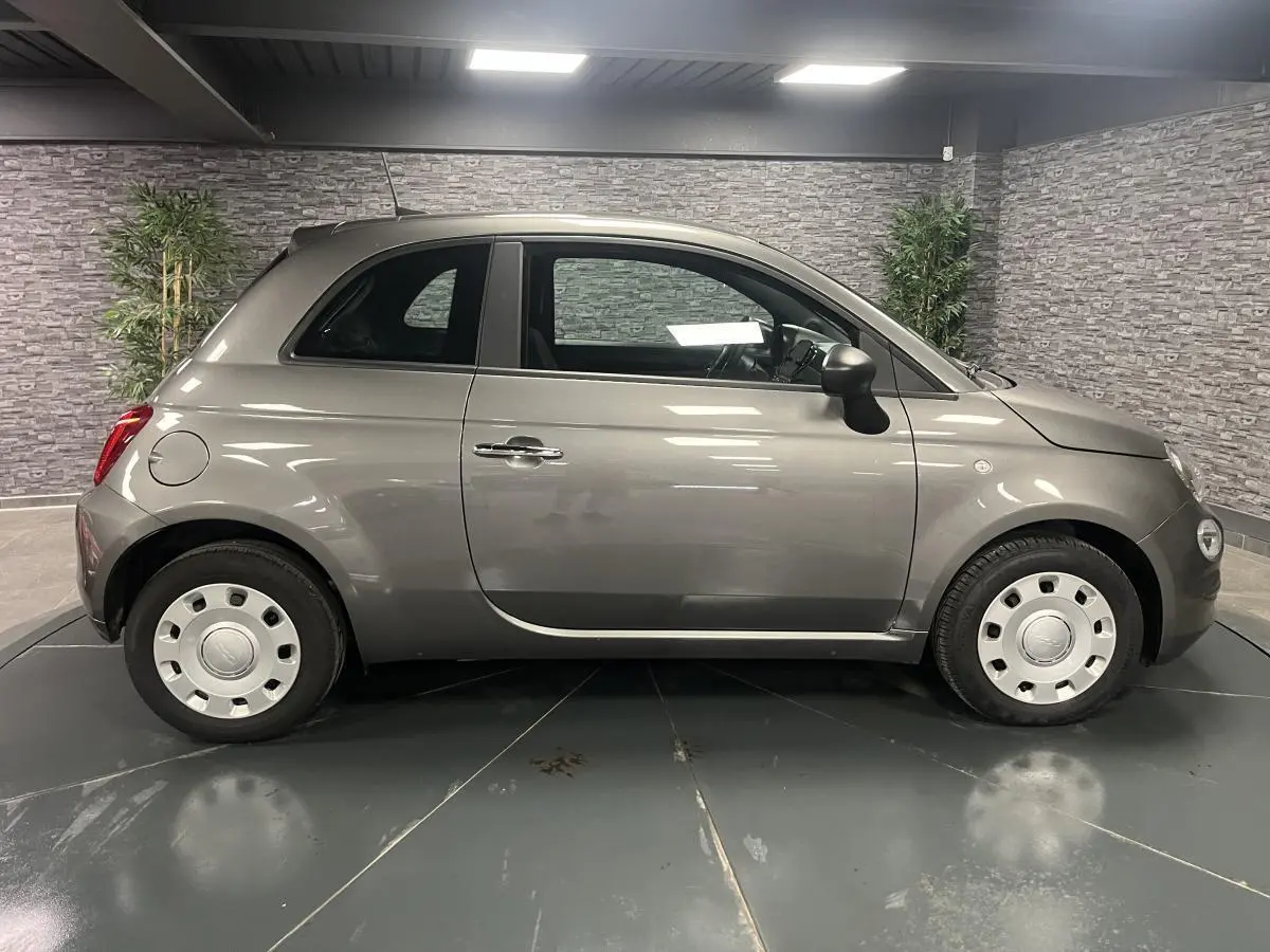 Profil droit d'une Fiat 500 gris métallisé 2023 avec jantes acier et poignées chromées en intérieur showroom.