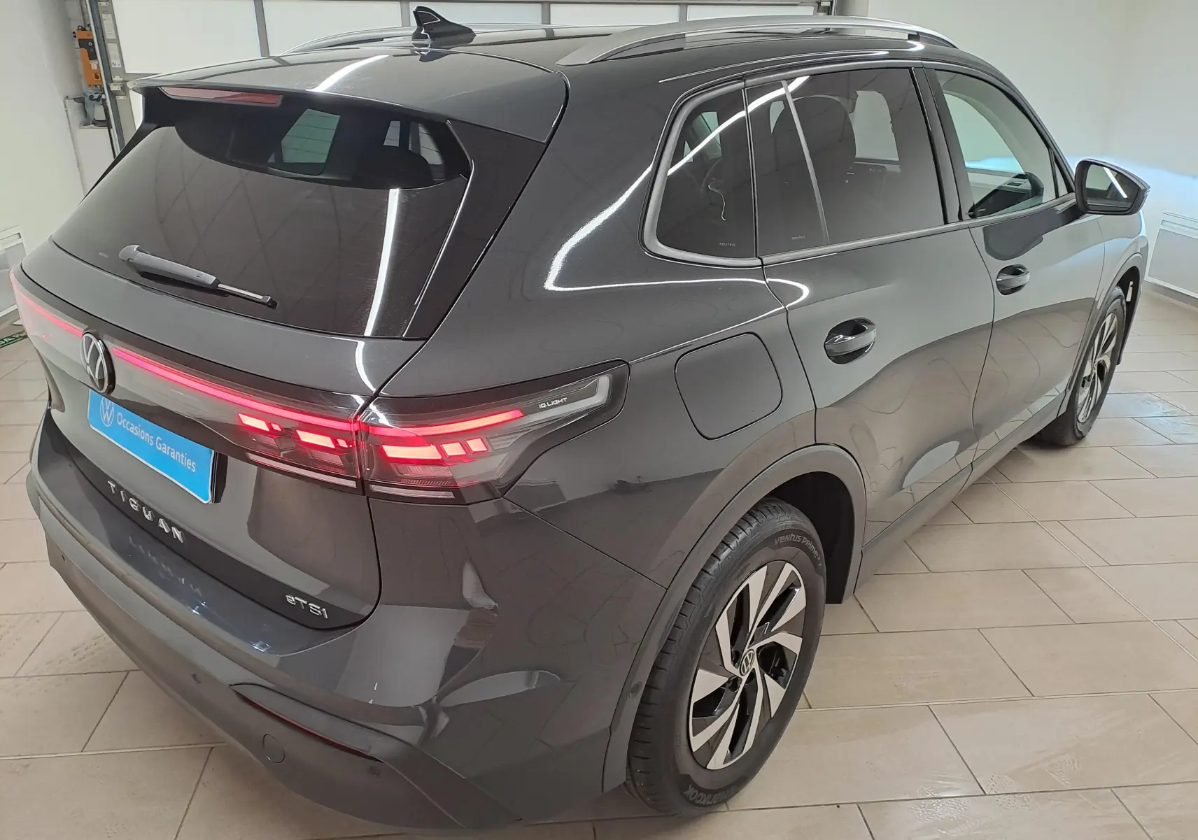 Vue 3/4 arrière droite du Volkswagen Tiguan 2024 gris Urano avec feux allumés et crochet d'attelage escamotable visible.