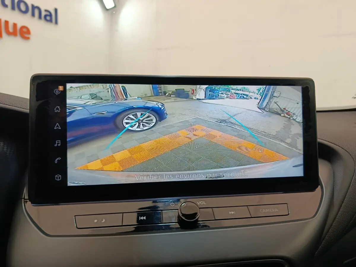 Écran tactile intérieur montrant la caméra de recul avec vue arrière sur un sol et une voiture bleue à gauche.