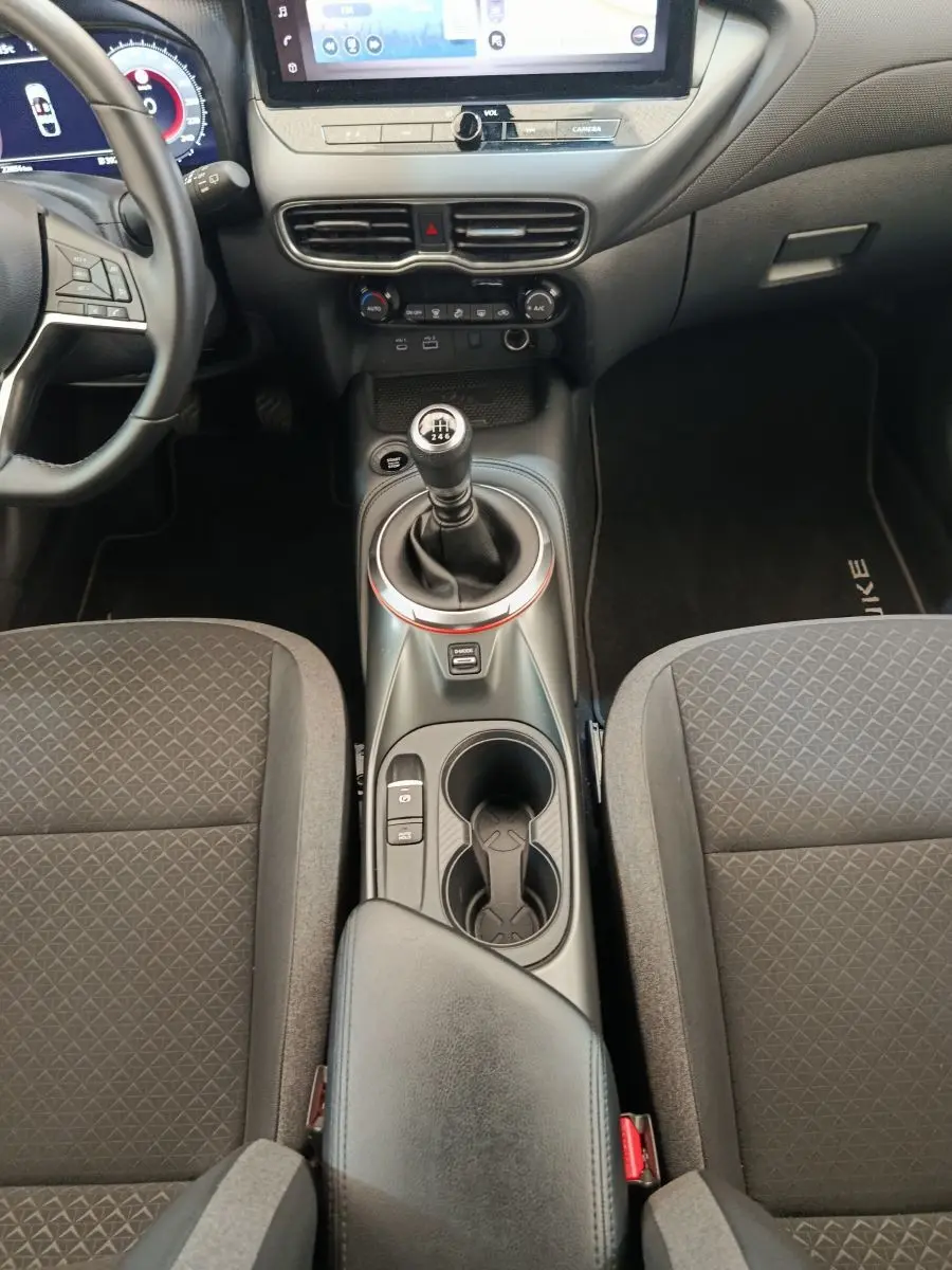 Vue intérieure centrée sur la console centrale et levier de vitesse manuel du Nissan Juke gris clair, avec accoudoir et sièges noirs.