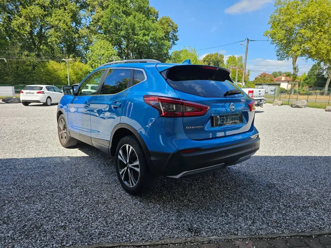 Nissan Qashqai bleu en 3/4 arrière droit, avec jantes alliage et toit ouvrant visible, stationné sur un sol gravillonné.