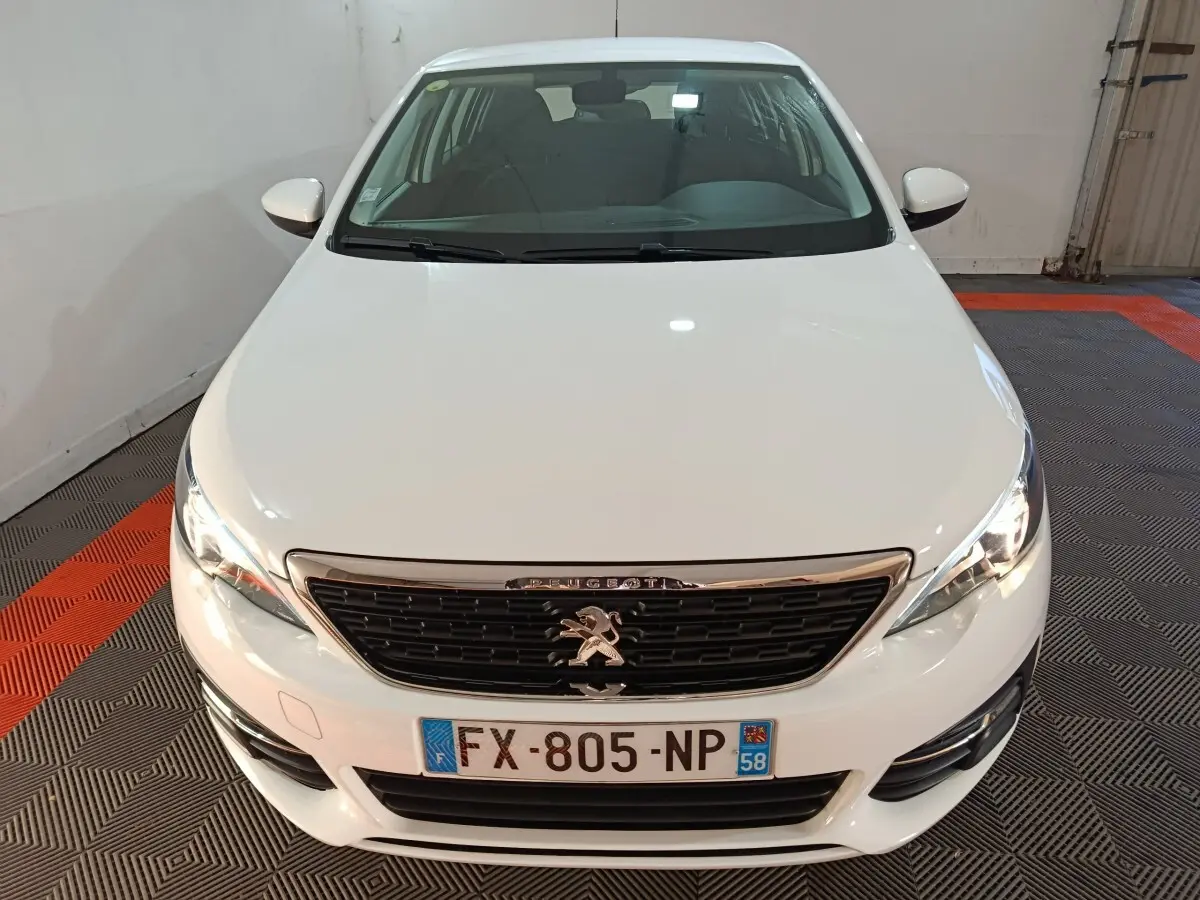 Vue avant d'une Peugeot 308 blanche 2021 avec calandre noire et phares allumés dans un garage.