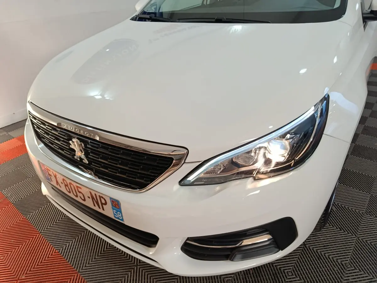 Vue 3/4 avant droit d'une Peugeot 308 blanche avec phares allumés et calandre chromée visible