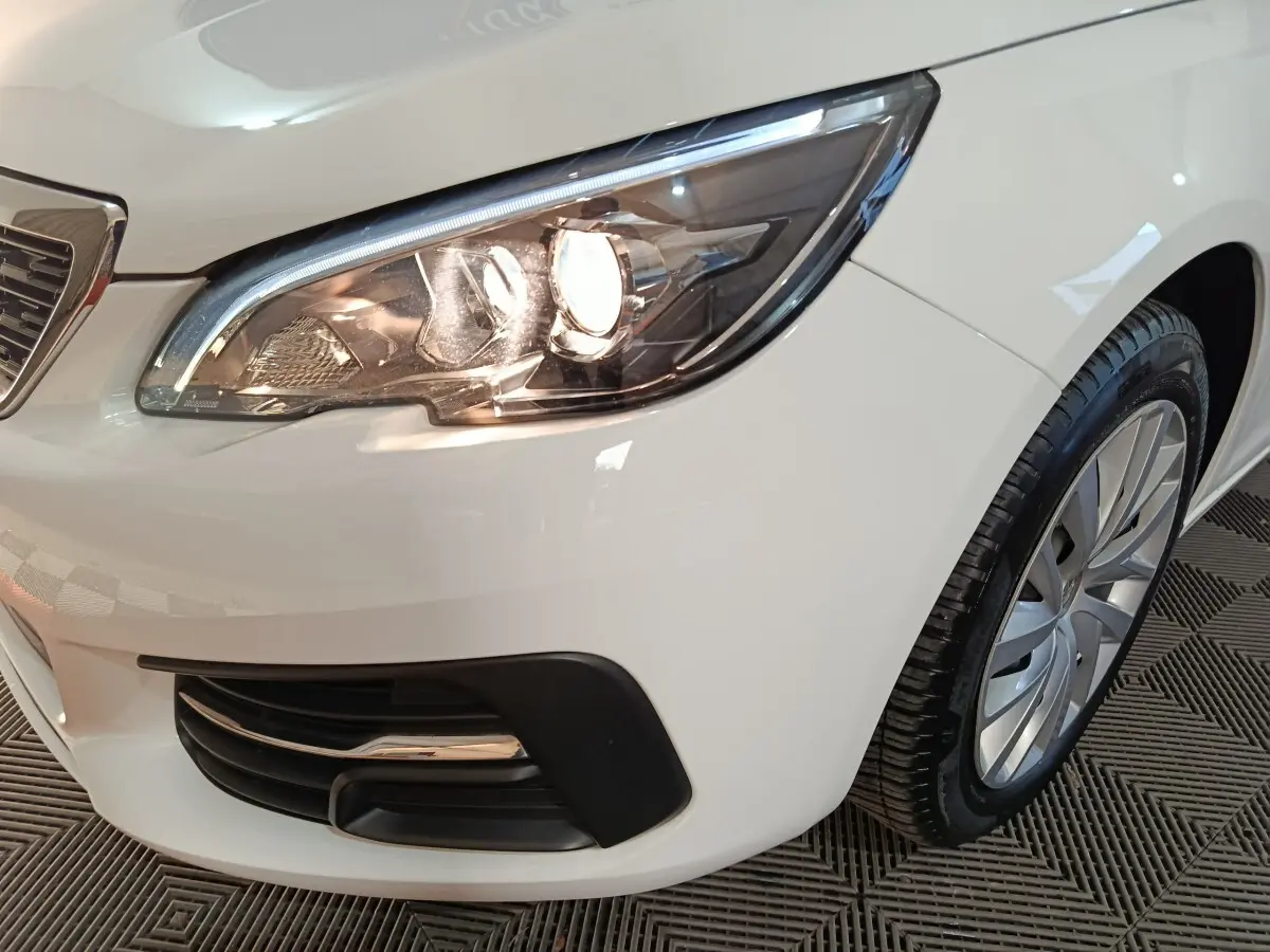 Gros plan sur le phare avant droit allumé d'une Peugeot 308 blanche, avec la roue et la calandre partiellement visibles.