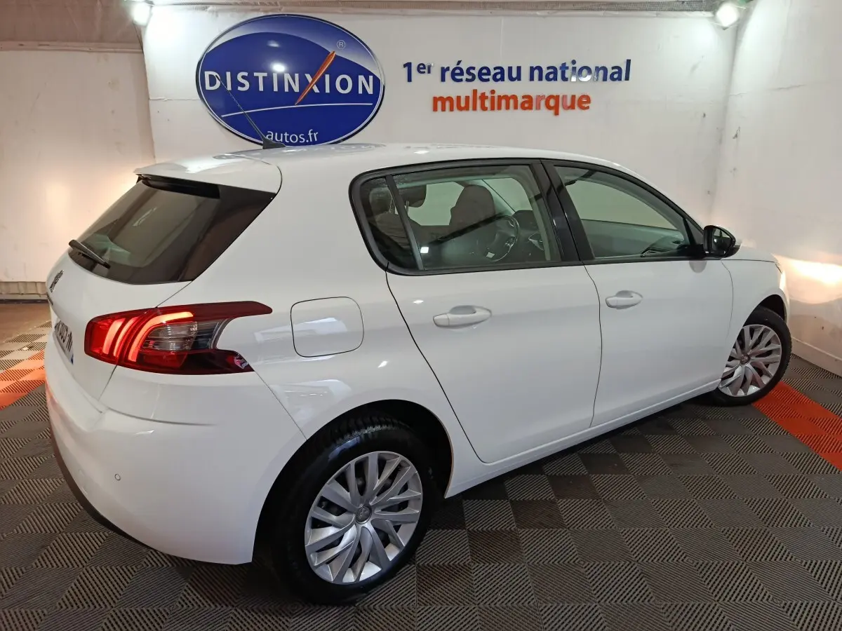 Peugeot 308 blanc vue 3/4 arrière droit en intérieur, feux arrière allumés et jantes alliage visibles