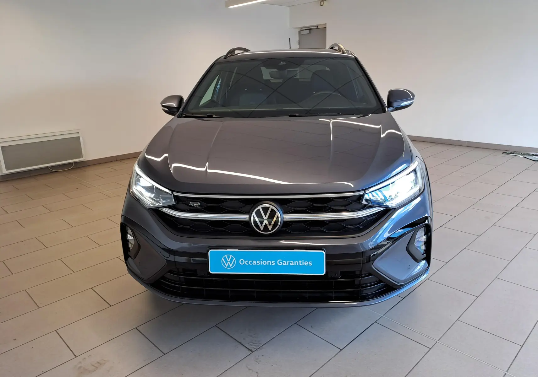 Vue avant d'un Volkswagen Taigo gris cendré 2025 avec calandre R-Line et phares allumés en intérieur showroom.