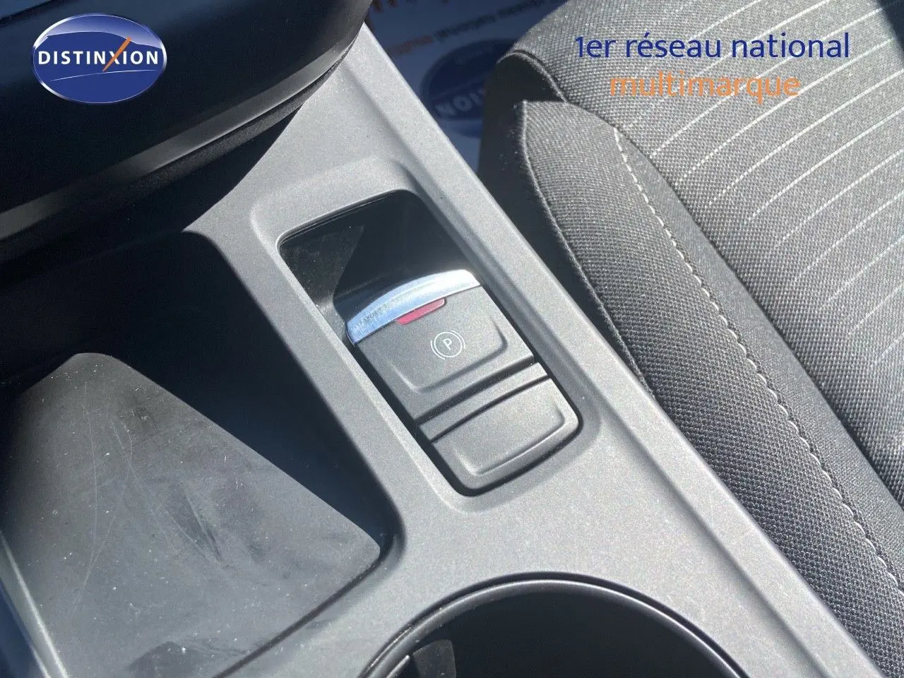 Gros plan sur la console centrale noire avec bouton de frein à main électronique et siège tissu gris à rayures.