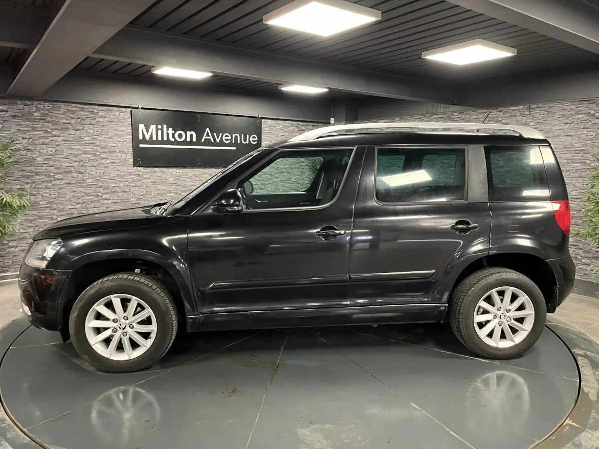 Profil côté gauche d'un Skoda Yeti noir 2015 avec jantes alliage 16'' et barres de toit argentées en intérieur showroom.