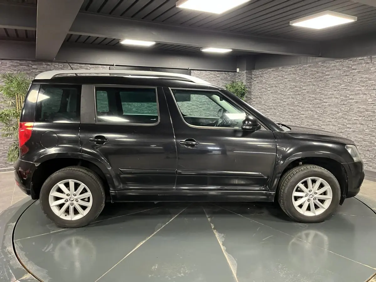 Profil côté gauche du Skoda Yeti noir 2015 avec jantes alliage 16 pouces et toit argenté en intérieur showroom.
