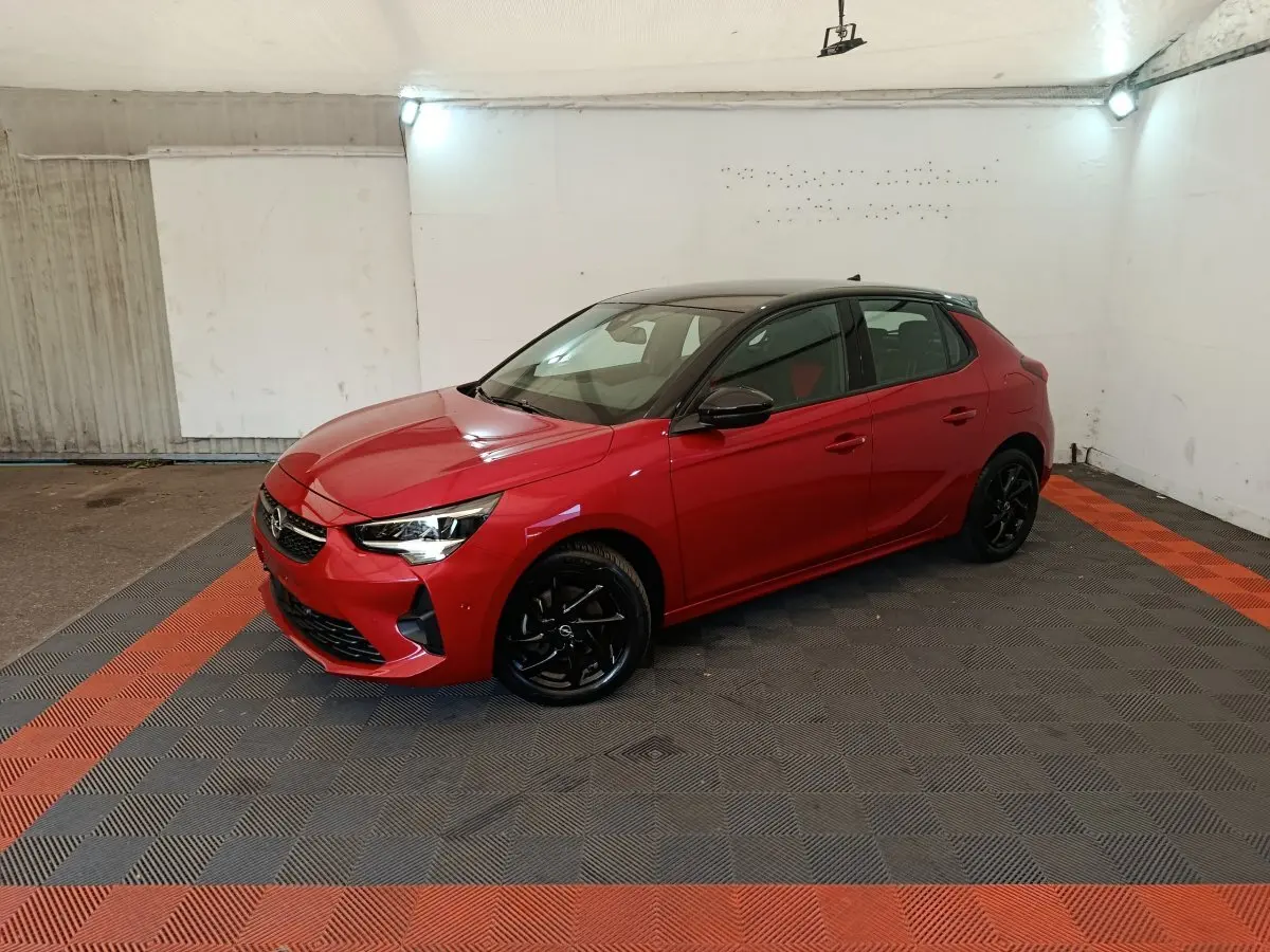Opel Corsa rouge 2023 vue 3/4 avant droit, toit noir, jantes alliage noires, dans un garage éclairé.