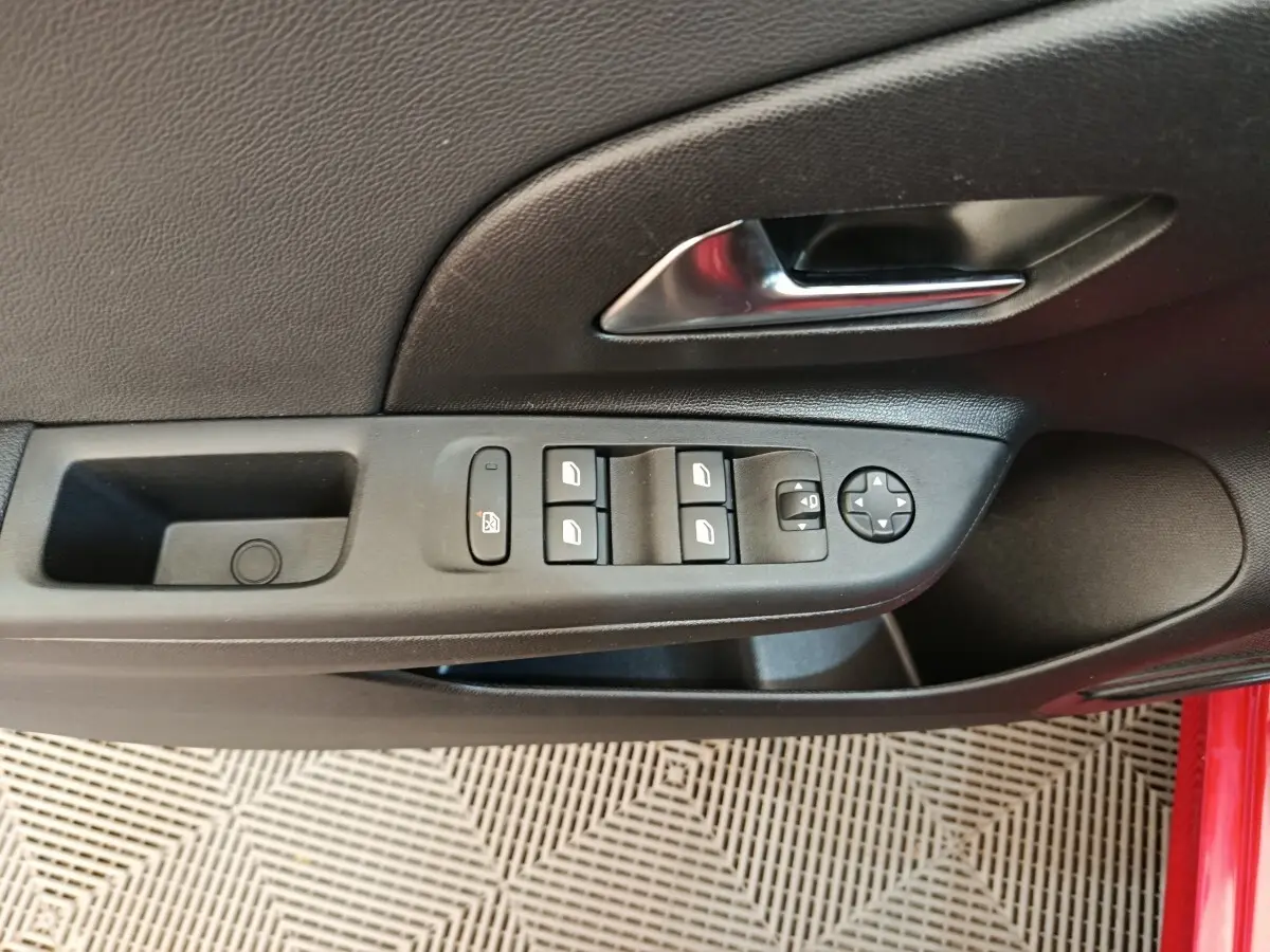 Vue rapprochée de la porte côté gauche de l’Opel Corsa rouge, avec commandes des vitres et poignée intérieure chromée.