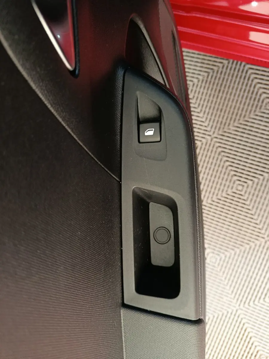 Détail de la commande de lève-vitre sur la porte intérieure noire d’une Opel Corsa rouge visible en arrière-plan.