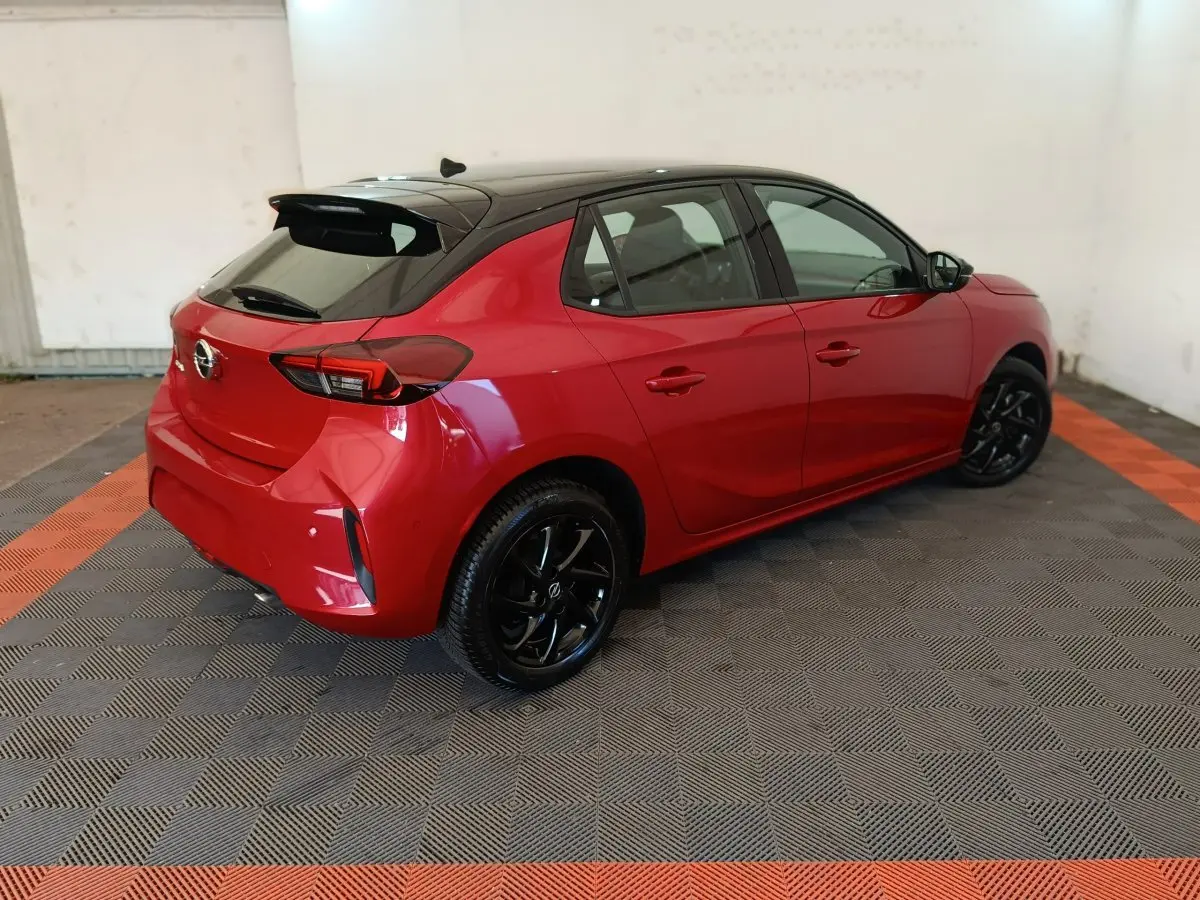 Opel Corsa rouge vue 3/4 arrière côté gauche, toit noir et jantes noires brillantes dans un garage.