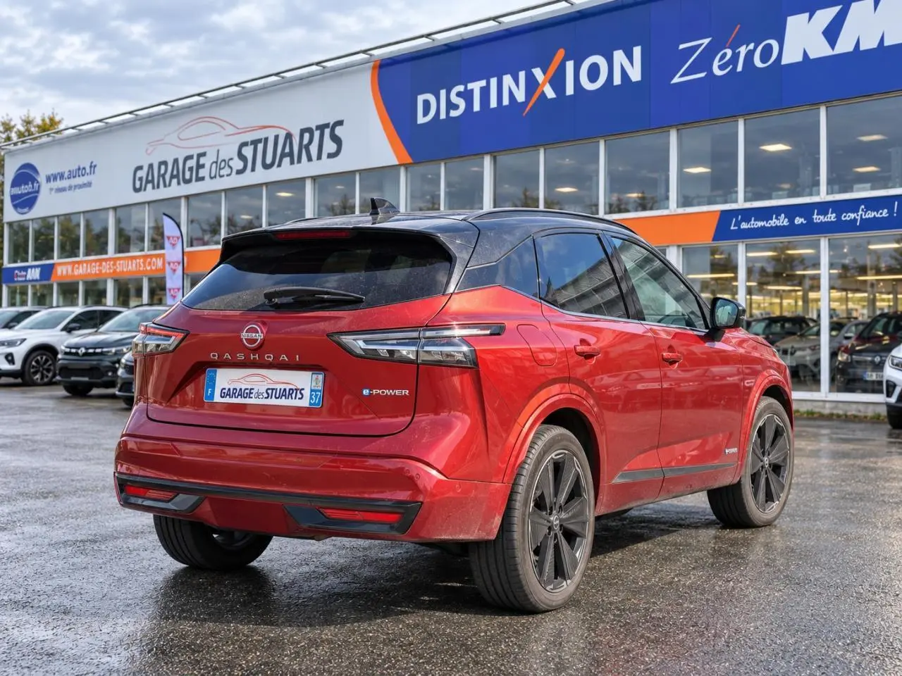 Vue 3/4 arrière d’un Nissan Qashqai 2024 rouge foncé e-Power Tekna garé devant un garage avec toit noir et jantes noires.