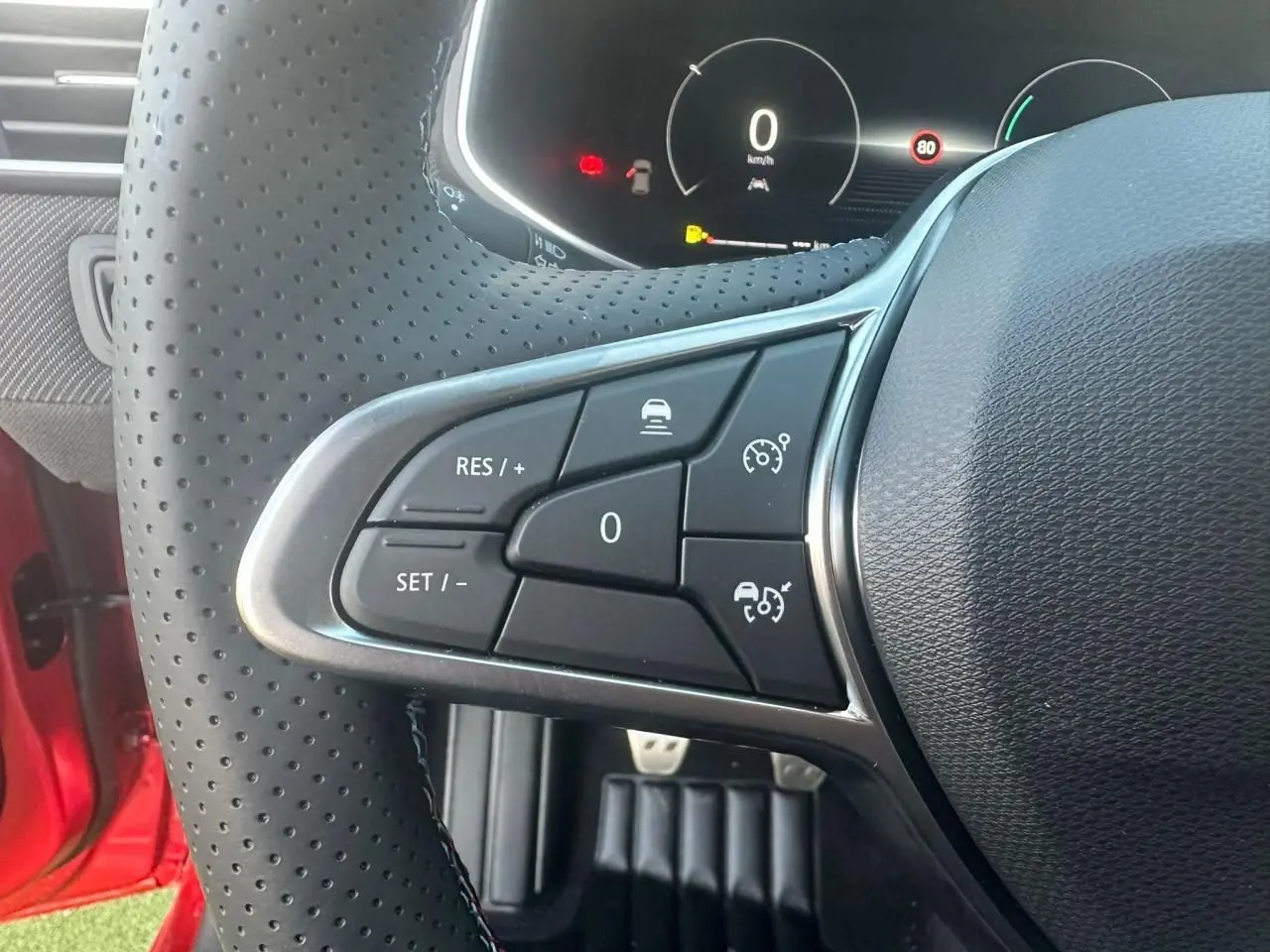 Gros plan sur les commandes au volant perforé de la Renault Clio V rouge flamme, affichage numérique en arrière-plan.