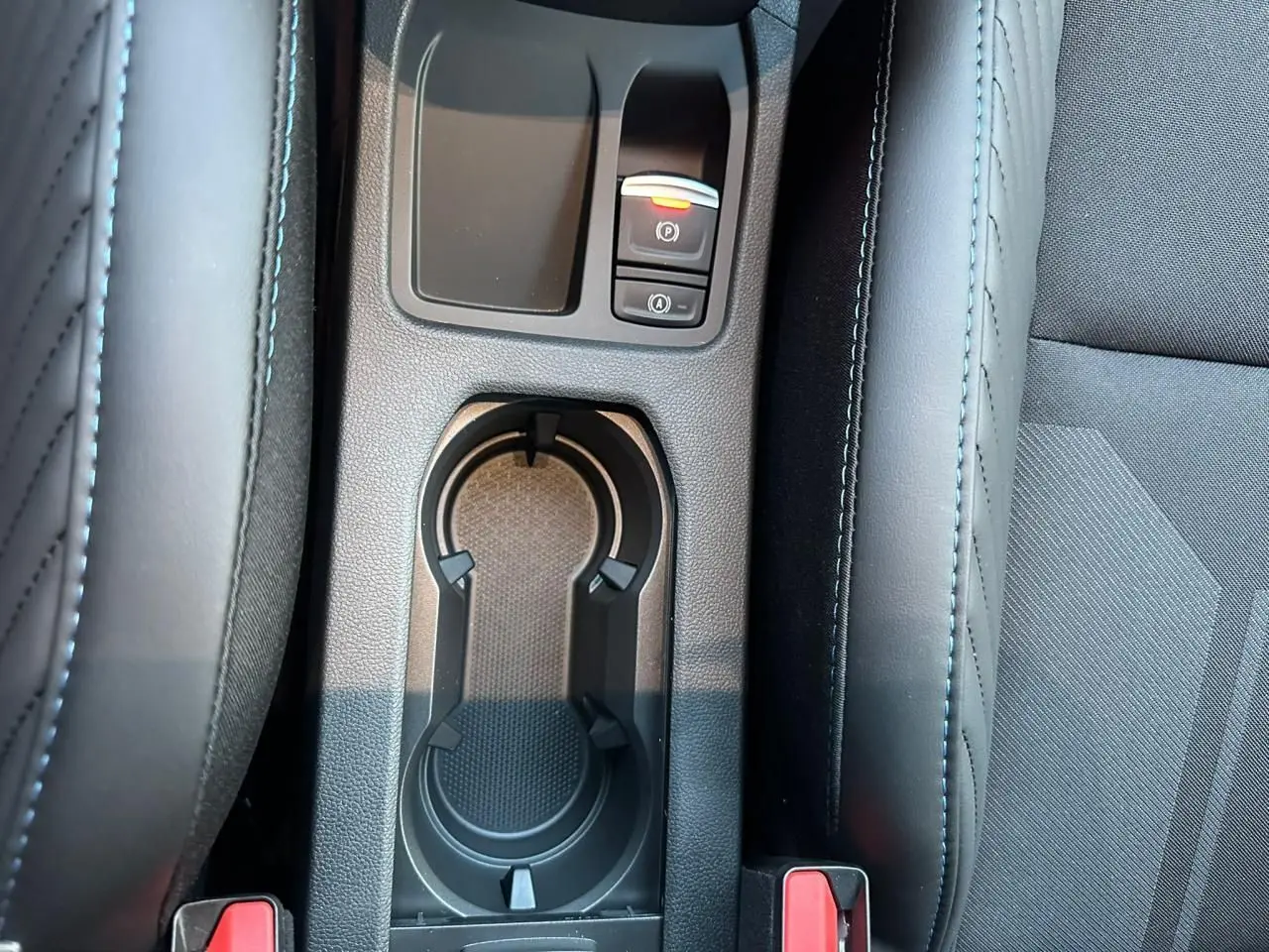 Gros plan sur la console centrale noire avec porte-gobelets et boutons de frein à main électrique dans une Renault Clio V rouge flamme.