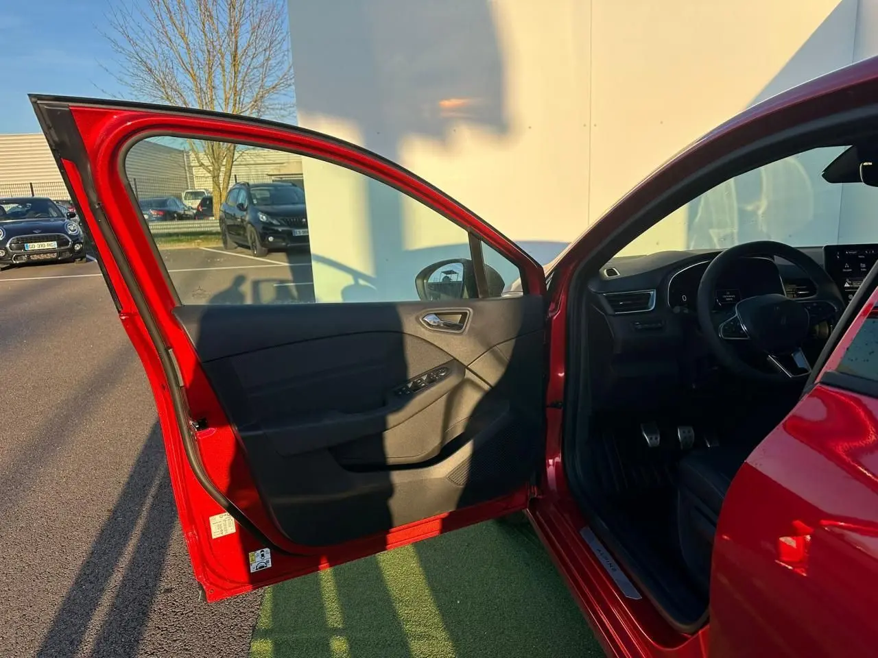 Vue côté gauche avant de la Renault Clio V rouge flamme avec porte ouverte et intérieur noir visible au soleil.