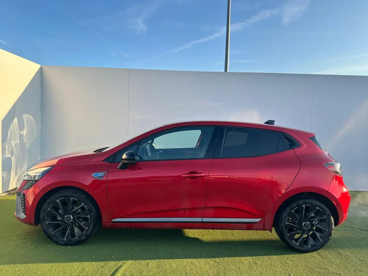 Profil côté gauche d'une Renault Clio V rouge flamme métal avec jantes alliage noires et antenne requin noire.