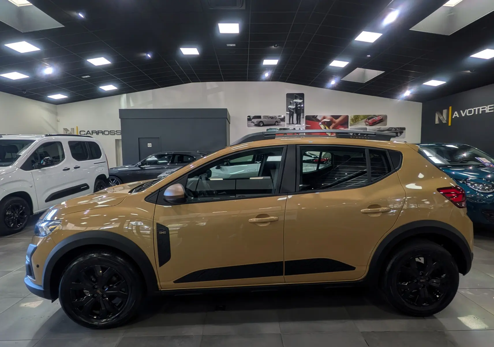 Vue latérale droite d'une Dacia Sandero Stepway beige Safari 2025 avec jantes noires et barres de toit en showroom.