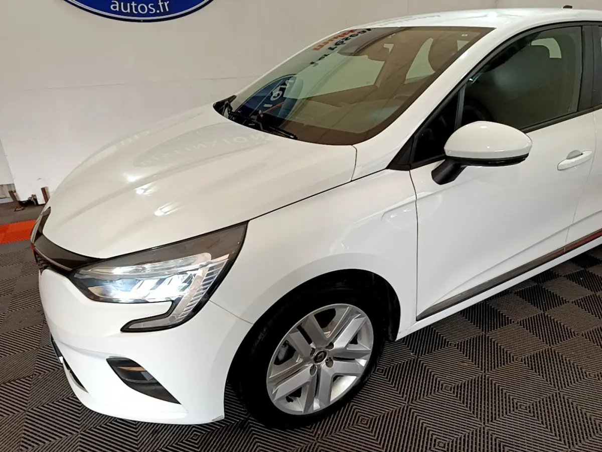 Vue 3/4 avant droit d'une Renault Clio Business Blue dCi 85 blanche avec phares LED allumés en intérieur showroom.