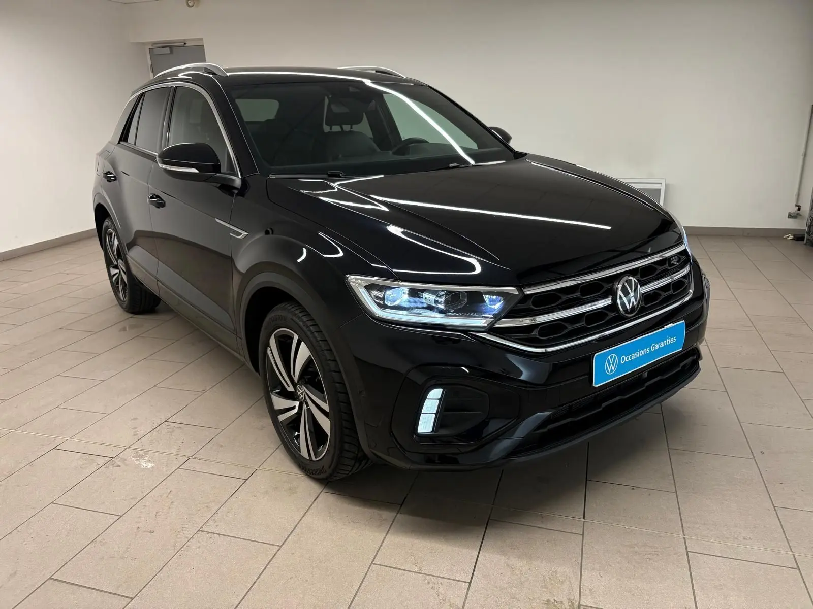 Volkswagen T-Roc noir intense en 3/4 avant droit, avec jantes alliage et phares LED allumés dans un intérieur neutre.