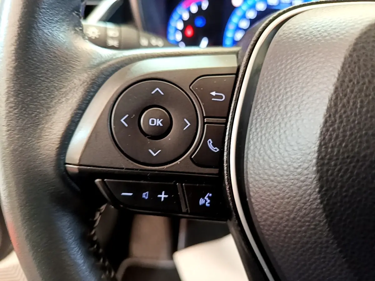 Gros plan sur les commandes noires du volant cuir de la Toyota Corolla hybride blanche, avec tableau de bord lumineux en arrière-plan.