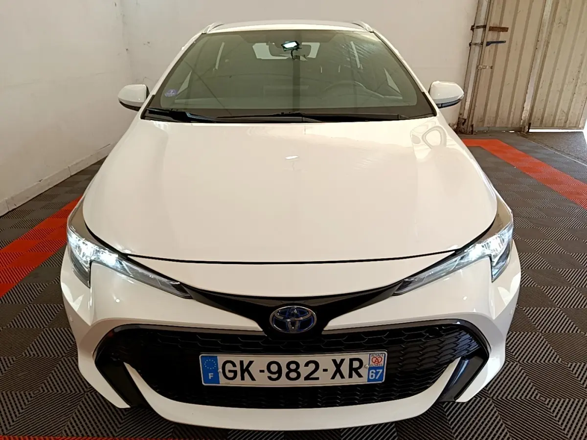Vue avant d'une Toyota Corolla hybride blanche avec calandre noire et phares LED allumés dans un garage.