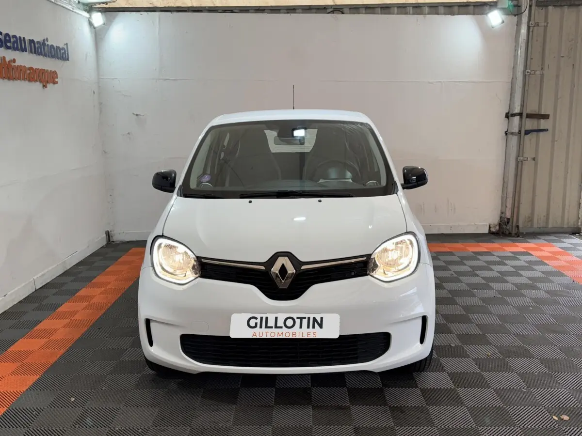 Renault Twingo blanc vue de face avec phares allumés dans un garage au sol gris et orange.