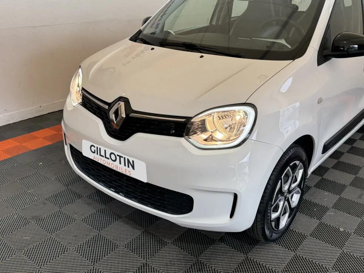 Vue 3/4 avant droite d'une Renault Twingo blanche 2022 avec phares allumés et jantes distinctives.