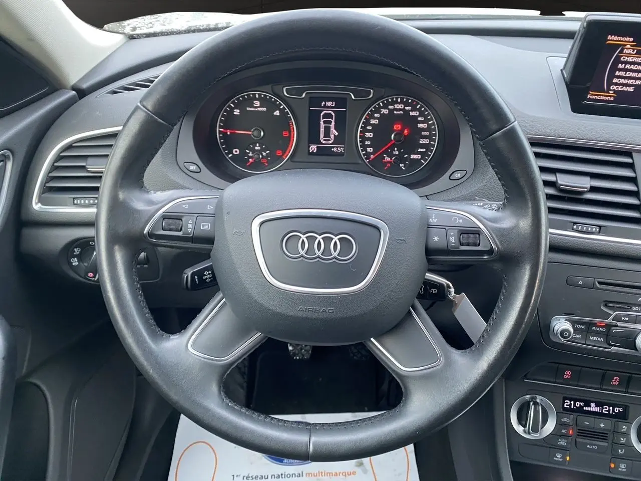 Vue intérieure centrée sur le volant cuir noir d’un Audi Q3 blanc de 2014 avec tableau de bord et écran GPS visible.