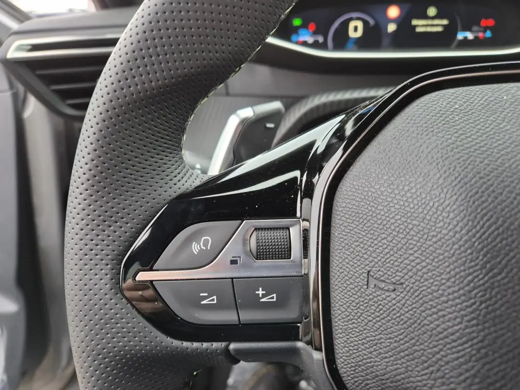 Gros plan sur le volant perforé noir de la Peugeot 208 Hybrid 110 e-DCS6 GT avec commandes intégrées et cockpit numérique en arrière-plan.