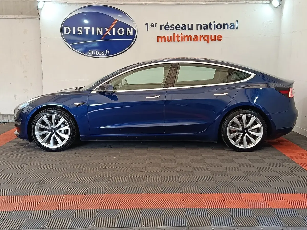 Profil droit d'une Tesla Model 3 Long Range bleu électrique, avec jantes argentées et lignes épurées visibles en intérieur showroom.