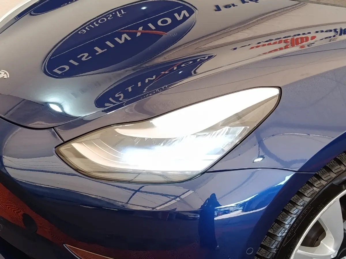 Gros plan sur le phare avant droit d’une Tesla Model 3 Long Range bleu électrique, avec reflet brillant sur la carrosserie.