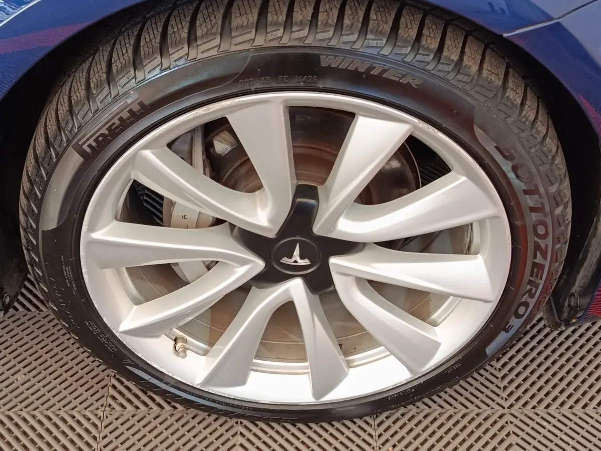 Gros plan sur la roue avant droite d'une Tesla Model 3 bleu, jante argentée avec logo Tesla au centre.