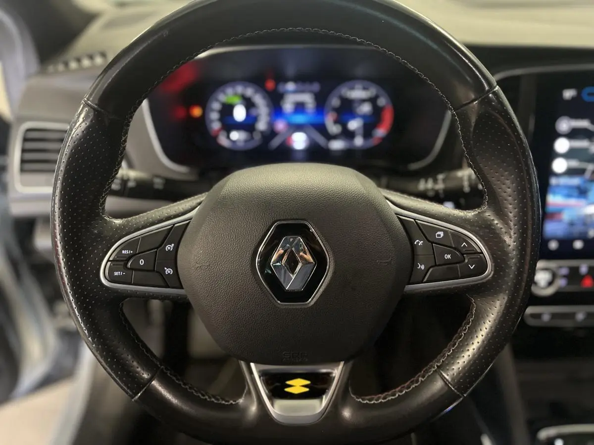Gros plan sur le volant cuir noir R.S. Line avec badge Renault, tableau de bord numérique flou en arrière-plan.