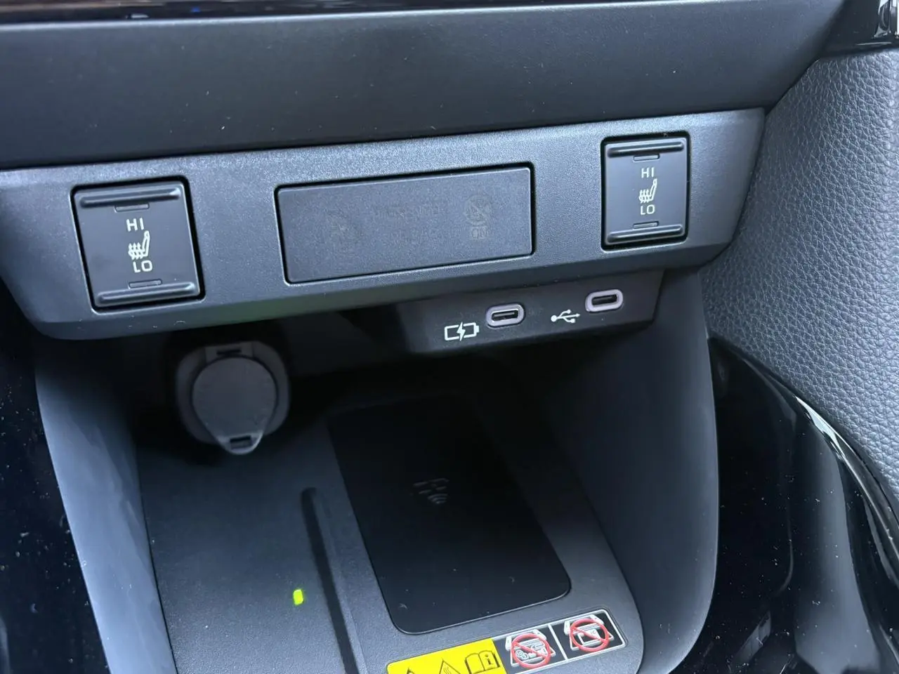 Gros plan sur la console centrale de la Toyota Yaris hybride 2025 montrant les commandes des sièges chauffants et un chargeur à induction noir.